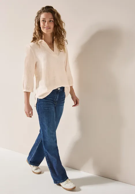 Frau mit lockigem Haar trägt helle Bluse, blaue Jeans und weiße Turnschuhe.